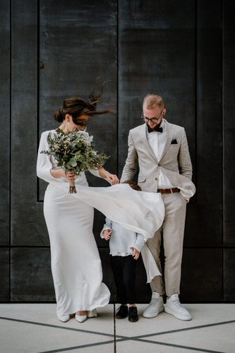 Hochzeitspaar steht mit Kind vor einer Wand, das wehende Kleid verdeckt das Gesicht des Kindes